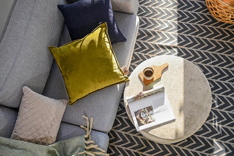 Een groen warmtekussen van bovenaf op een grijze bank met andere sierkussens erbij.